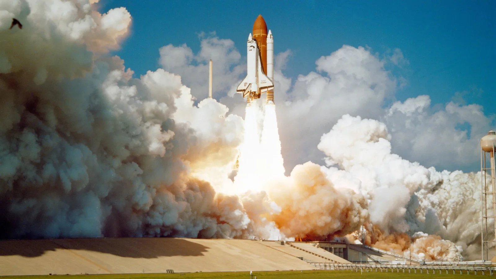 Space Shuttle Challenger lifting off from Kennedy Space Center, engulfed in launch smoke.